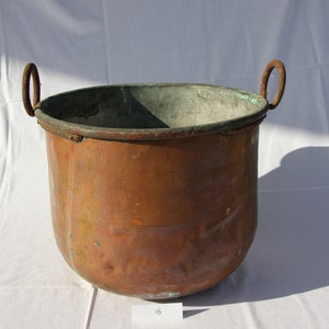 May include: A large, copper pot with two iron handles. The pot has a rounded bottom and a wide, open top. The copper has a patina of age and use.