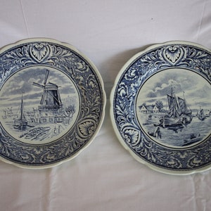 Puede incluir: Dos platos de cerámica azul y blanco con bordes decorativos. Un plato presenta una escena de molino de viento, mientras que el otro representa un velero.