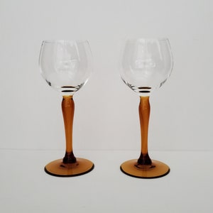 May include: Two clear glass wine glasses with amber colored stems and bases. The glasses are empty and sitting on a white surface.