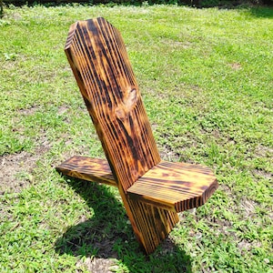 Könnte beinhalten: Ein rustikaler Holzstuhl mit einer gebrannten Oberfläche. Der Stuhl hat ein einfaches Design mit einer breiten Rückenlehne und einer Sitzfläche, die sich zur Seite erstreckt. Der Stuhl steht auf einem Rasen.