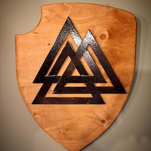 Può includere: Scudo in legno con un simbolo Valknut in metallo scuro. Lo scudo è di colore marrone chiaro e a forma di scudo. Il simbolo Valknut è composto da tre triangoli interconnessi.