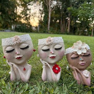 May include: Three decorative face planters with closed eyes, red lips, and lace headbands. The planters are in shades of beige and brown, adorned with floral accents and jewelry. They are placed on a bed of green grass.