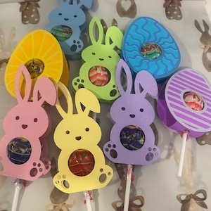 May include: A collection of colorful Easter-themed lollipop holders. The holders are shaped like bunnies and eggs in various pastel colors, each holding a lollipop. The background features a repeating pattern of bunny illustrations.