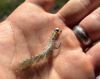 May include: A handmade fishing fly with a silver bead head and a curved hook. The fly features a body of brown, green, and white fibers, with iridescent accents. This fly is designed for catching fish.