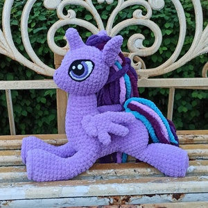 May include: Purple crocheted pony toy with a blue and purple mane sitting on a white metal bench.