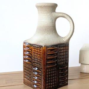 May include: A square-shaped ceramic pitcher featuring a textured, glossy brown glaze on the lower body with alternating panels of raised oval and grid patterns. The upper neck and handle are finished in a matte, speckled off-white glaze.