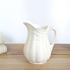 May include: Off-white ceramic pitcher with a handle and a decorative floral design. The pitcher has a rounded body with vertical ridges and a curved spout. It is sitting on a wooden surface.