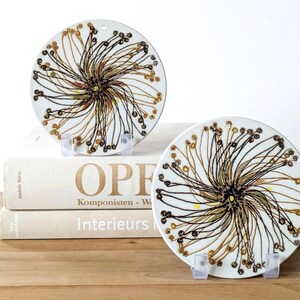 May include: Two white ceramic decorative plates with a floral design in brown, black, and yellow. The plates are displayed on clear stands, resting on a stack of books with the word "OPERA" visible. The plates have a small hole at the top.