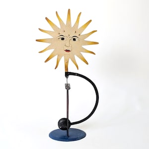 Könnte beinhalten: Eine dekorative Metallskulptur mit einem Sonnengesicht, gelb getönten Strahlen, dunklen Augen und roten Lippen. Die Sonne ist an einem schwarzen, gebogenen Arm befestigt, der auf einem blauen, runden Sockel steht, mit einer kleinen schwarzen Kugel.