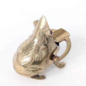 Puede incluir: Un cenicero de latón con forma de rana con patrones florales grabados. El cenicero tiene un pequeño asa y una ranura para cigarrillos. Una pequeña joya roja está engastada en la cabeza de la rana.