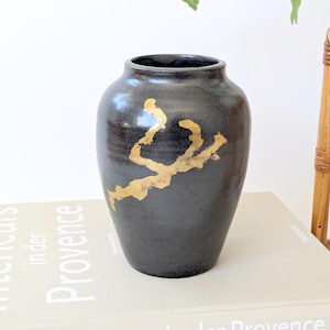 Pode incluir: Vaso de cerâmica preta com um design abstrato dourado. O vaso tem formato arredondado e uma abertura larga. Está sobre um livro com o título "Interieurs in der Provence".