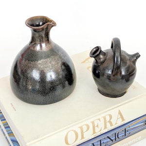 May include: Two ceramic vessels with a dark, speckled glaze. One is a rounded vase with a spout, the other a small pitcher with a handle. They sit atop a stack of books with gold lettering.