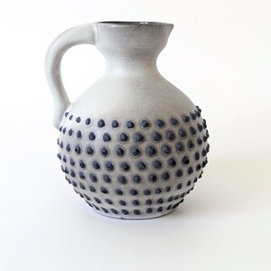 Può includere: Una brocca in ceramica con corpo arrotondato e manico curvo. La superficie è di colore bianco sporco opaco, caratterizzata da un motivo strutturato di piccoli punti blu scuro in rilievo che coprono i due terzi inferiori del recipiente.