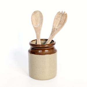 May include: A ceramic kitchen utensil holder with a brown rim and base, and a tan body. Two wooden utensils, a spoon and a fork, are placed inside the holder. The background is white.