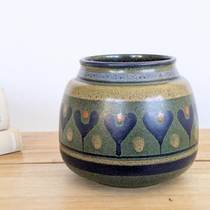 May include: A round ceramic vase with a speckled olive green finish, featuring a horizontal band of dark blue heart-shaped motifs and small orange dots. The vase has a wide, short neck with a dark blue rim and a solid dark blue base stripe.