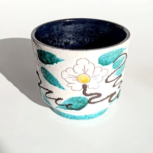 Peut inclure: Une tasse en céramique à base blanche, ornée d'un motif floral avec une fleur blanche au centre jaune et des feuilles turquoise. Le bord est bleu foncé et la tasse a une bande turquoise à la base. Des lignes noires soulignent le motif.