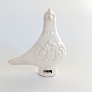 Puede incluir: Figurita de cerámica blanca en forma de paloma con detalles florales en relieve. La paloma tiene un pequeño pico y un cuerpo redondo. La figurita mide aproximadamente 25 cm de alto y tiene una etiqueta negra en la parte inferior que dice "Rain Song".