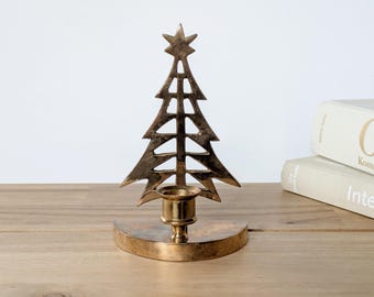 May include: A gold-coloured Christmas tree candle holder with a star on top. The tree-shaped design has cut-out details and a round base. The candle holder is on a wooden surface.