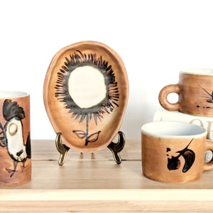 May include: A collection of handmade ceramic tableware with a rustic aesthetic. The set includes a tall tumbler with a rooster design, a sunflower-shaped platter, and two mugs with abstract black designs. The pieces are brown with white interiors.
