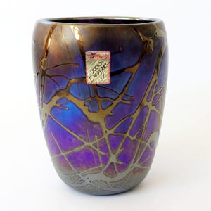 Puede incluir: Un jarrón de vidrio decorativo con un diseño único. El jarrón presenta una mezcla de colores, incluyendo morado, azul y dorado, con una red de líneas doradas. La etiqueta dice "ASKA Studio Design".