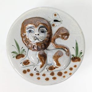 Peut inclure: Assiette en céramique avec un motif de lion en relief. Le lion a une crinière et des taches marron, avec un papillon et des accents de feuillage. L'assiette est ronde avec un fond bleu clair et des détails marron et vert.