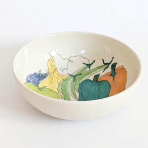 May include: Cream-colored ceramic bowl with a hand-painted vegetable design. The bowl features illustrations of a blue onion, yellow squash, green beans, green and orange bell peppers. The bowl is set against a white background.