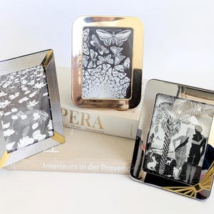 Puede incluir: Tres marcos de fotos plateados con detalles dorados. Cada marco contiene una imagen en blanco y negro. Uno presenta mariposas, otro una escena natural y el tercero una escena con figuras y perros.