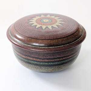 May include: A round, lidded ceramic container with a sunburst design on the lid. The lid and base are primarily brown with horizontal bands of color. The sunburst has yellow, blue, and orange accents.
