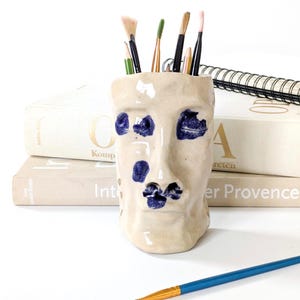 Pode incluir: Um porta-lápis de cerâmica na cor creme com formato de rosto humano e traços texturizados. O rosto apresenta detalhes esmaltados em azul escuro nos olhos e na boca. Vários pincéis e lápis estão guardados dentro da abertura superior.