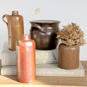 May include: A collection of antique stoneware pottery pieces. Includes a brown jug with a handle, a dark brown pot with handles, a small brown vase with dried flowers, and a reddish-brown bottle. The items are arranged on books.