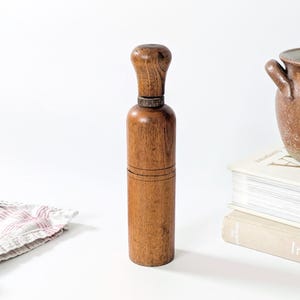 Op de afbeelding: Een hoge, cilindrische houten pepermolen met een afgeronde handgreep aan de bovenkant en een zichtbare houtnerf. De molen heeft een donkere metalen ring die de handgreep van het hoofdgedeelte scheidt, met een verticaal ontwerp.