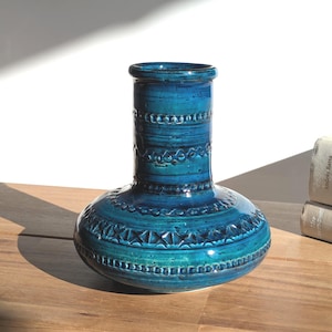 Op de afbeelding: Een turquoise keramische vaas met een bolle basis en een cilindrische hals. De vaas heeft een getextureerd, patroonontwerp met donkerblauwe accenten. De vaas staat op een houten oppervlak, met zonlicht dat een schaduw werpt.