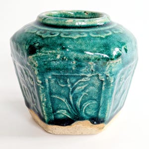 Könnte beinhalten: Eine vintage, teal-farbene Keramikvase mit breiter Öffnung und strukturiertem, facettiertem Körper. Die Vase weist geprägte Blumenmuster und einen hellfarbigen Sockel auf. Die Glasur hat ein glänzendes Finish.