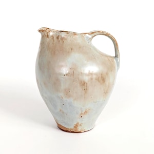 Può includere: Brocca in ceramica fatta a mano con manico. La brocca ha una combinazione di colori beige chiaro e blu con accenti marroni. Ha un beccuccio per versare liquidi e viene probabilmente utilizzata per servire acqua o altre bevande.
