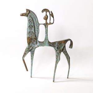 Könnte beinhalten: Eine bronzefarbene Metallskulptur eines stilisierten Pferdes mit Reiter. Die Figur weist eine verwitterte, blau-grüne Oxidationspatina auf, mit komplexen geometrischen Mustern, die in die Oberfläche des Pferdekörpers und der Reiterform eingraviert sind.