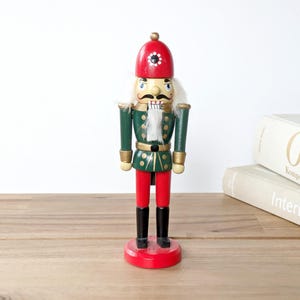 May include: A wooden nutcracker soldier figurine. The figurine has a red hat, green jacket, red pants, and black boots. The nutcracker has a white beard and mustache. The figurine is standing on a red base. The background includes two books and a wooden surface.