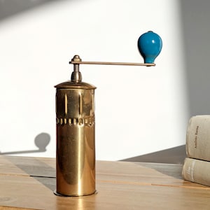 以下が含まれることがあります： 青いハンドルが付いたヴィンテージの真鍮製コーヒーグラインダー。グラインダーは、底の近くに一連の小さな穴がある円筒形のボディをしています。ハンドルは細長いアームに取り付けられています。グラインダーは木製の表面にあります。高さ約20cm。
