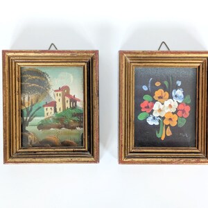 May include: Two framed miniature paintings. One depicts a landscape with a building and a tree. The other shows a bouquet of flowers against a black background. Both have gold-colored frames with a red inner edge.