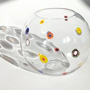 May include: A clear, round glass bowl with colorful floral designs. The bowl is decorated with various small, circular patterns in shades of blue, yellow, red, and green. The bowl casts a shadow on a white surface.