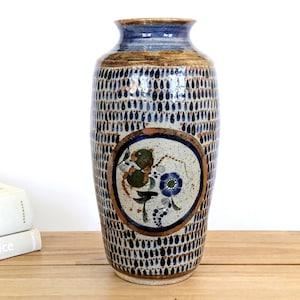 Op de afbeelding: Een hoge keramische vaas met een gespikkelde crèmekleurige basis met een herhalend patroon van kleine donkerblauwe druppelvormen. Het midden toont een cirkelvormig medaillon met een handgeschilderd bloemmotief in blauw-, groen- en bruintinten.