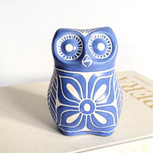 Może przedstawiać: Niebieska ceramiczna figurka sowy z białymi wzorami kwiatowymi. Sowa ma duże, okrągłe oczy i stylizowany design. Figurka stoi na książce z widocznym na okładce słowem "OPERA".