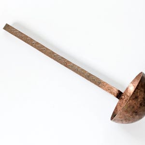 Op de afbeelding: Een koperen pollepel met een lange, platte steel en een afgeronde kom. De steel is versierd met een herhalend patroon van kleine, hartvormige ontwerpen. De pollepel heeft een rustieke, antieke uitstraling.