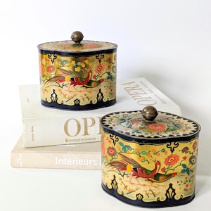 May include: Two oval metal tins with lids, each featuring a gold background, black trim, and a colourful illustration of two pheasants surrounded by floral patterns. Each lid is topped with a small, round, metallic knob for easy opening.