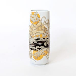Può includere: Un vaso in ceramica bianco, alto e rettangolare, con un motivo decorativo. Il design presenta cerchi gialli, forme a nuvola dorate e grigie e una striscia orizzontale nera. Un elemento decorativo per la casa.