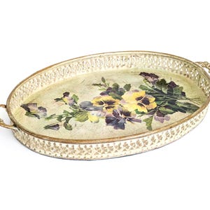 Könnte beinhalten: Ein ovales Metalltablett mit cremefarbener Antik-Optik und goldfarbenen Rändern. Das Tablett verfügt über einen dekorativen Gitterrand, zwei verzierte Griffe und eine zentrale Blumenillustration mit gelben und violetten Stiefmütterchen.
