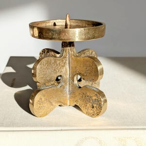 Könnte beinhalten: Ein Vintage-Kerzenhalter aus Messing mit einem einzigartigen, skulpturalen Design. Der Kerzenhalter hat eine runde Oberseite für die Kerze und einen dekorativen Sockel. Das Messing hat eine warme, gealterte Patina, die seinen antiken Charme unterstreicht.