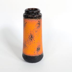 May include: A cylindrical ceramic vase featuring a vibrant orange body with a mottled black texture. The top rim and the base are finished in a dark, solid black glaze, creating a distinct contrast against the bright orange centre section.