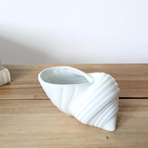 Peut inclure: Un vase blanc en céramique en forme de coquillage avec un extérieur texturé et strié. Le vase a un intérieur lisse et ouvert et repose sur une surface en bois. Le design rappelle la décoration côtière.