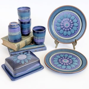 May include: A collection of blue and purple ceramic tableware, including plates, cups, and a butter dish. The plates feature a floral design in shades of blue and purple. The cups and butter dish have a striped pattern. The items are arranged on a white surface.