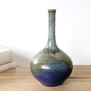 May include: A handcrafted ceramic vase with a bulbous base and a slender neck. The vase features a gradient of colors, transitioning from a deep blue at the base to olive green and then to a lighter blue at the top. It stands on a wooden surface.